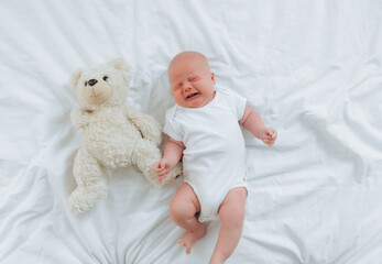 a 2-month-old baby is crying in bed with a teddy bear. top view