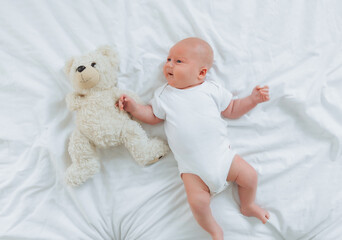 a 2-month-old baby in a white bodysuit is lying at home on a white bed next to a teddy bear. view from above. newborn