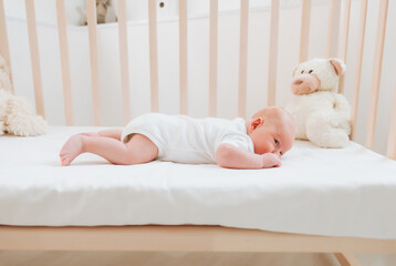 A charming smiling blue-eyed 2-month-old baby in a white bodysuit lies in a crib next to a teddy...