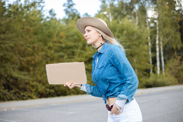 Young woman hopefully look out passing cars with empty cardboard poster on roadside in forest. Lady...