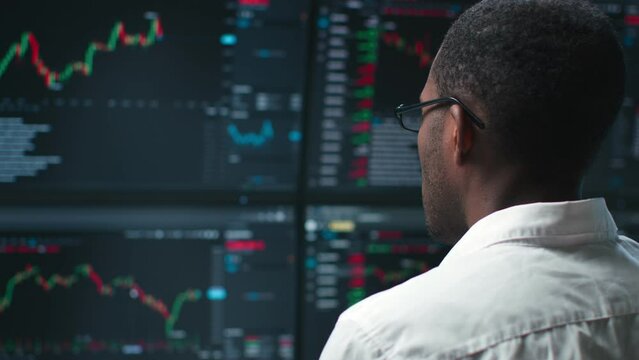 Back view of African-American broker looking at multi-screens with stock market data