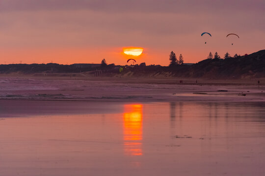 A colourful sunset reflecting on a wet beach with parasailers in the sky