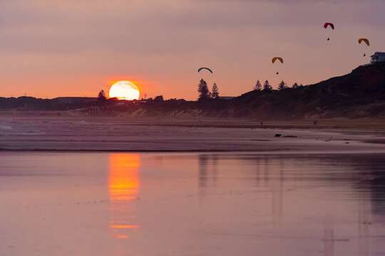 A colourful sunset reflecting on a wet beach with parasailers in the sky