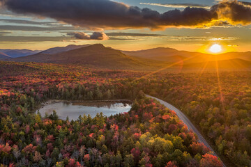 Sunrise over a scene of a road and lake through a forest with trees in fall colors of red, orange...
