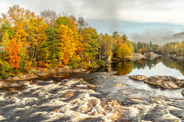 A river transitioning from mirror calm to rapids over rocks, the shores are lined with trees in full fall colors