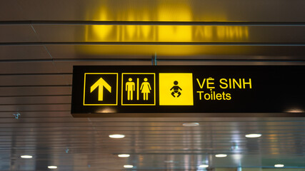 Toilets with arrow and gender icon  lighting signboard (English and Vietnamese text) which is installed on ceiling of the airport terminal building, selective focus.