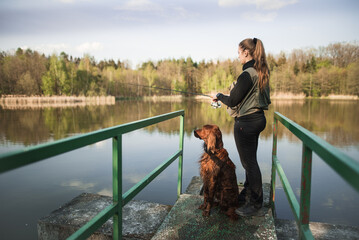 A young nice woman in a vest is fishing on a small pond with her hunting dog. Catching fish in nature at sunset. A relaxing hobby. 