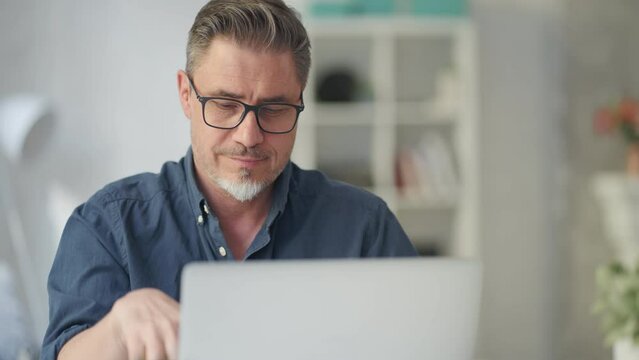 Older man sitting at desk in bright room working on laptop computer in home office. Mature age, middle age, mid adult casual man in 50s, confident happy smiling.