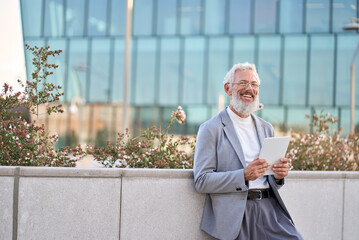 Happy european bearded stylish old mature adult professional business man, smiling senior older businessman ceo holding digital tablet using fintech tab standing outside big city office buildings. - obrazy, fototapety, plakaty