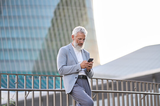 Stylish Classy Older Mature Adult Professional Business Man, Senior Old Bearded Businessman Executive Holding Smartphone, Using Mobile Tech On Cell Phone Standing Outside Big City Office Buildings.
