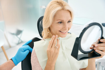 Happy smiling woman is looking to the mirror in dental chair
