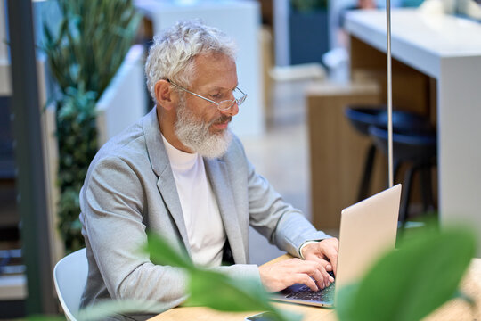 Busy Smart Older Senior Professional Business Man Wearing Spectacles Using Laptop Sitting In Corporate Office At Desk. Middle Aged Old Adult Concentrated Businessman Working Online Typing On Computer.