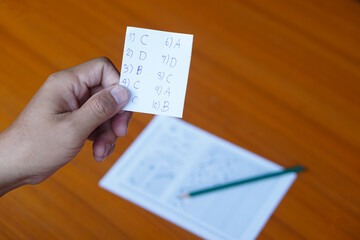 Naklejka premium Closeup student's hand holds small piece of paper with answers during doing the test on pape . Concept : Cheat the test. Dishonest behaviour. Education assessment. 