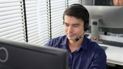 Young competent asian male call center agent working at his computer while simultaneously speaking...