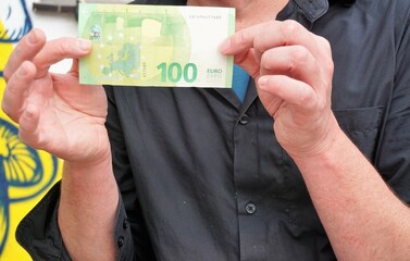 Mann in grauem Hemd hält 100 Euro Schein mit seinen Händen vor Brust 