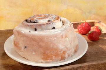 Freshly baked bun with white icing and pieces of chocolate chips on a white plate on a yellow background with fresh ripe strawberries