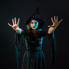 A charming young witch in a costume makes magical passes with two hands, a caster. Young brunette in pointed hat and black dress, photo on black