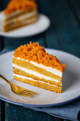 Carrot cake with cream cheese in layers and carrots on top on a  blue and white background with carrots in blurry background