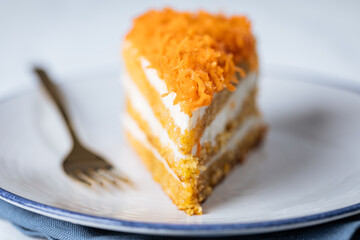 Carrot cake with cream cheese in layers and carrots on top on a  blue and white background with carrots in blurry background