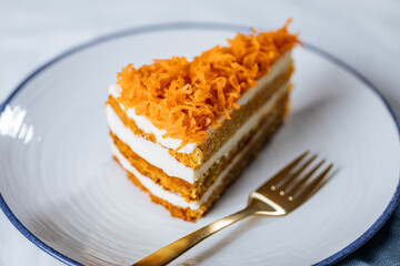 Carrot cake with cream cheese in layers and carrots on top on a  blue and white background with carrots in blurry background