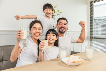 Fototapeta premium Asian family enjoying breakfast at living room. little girl daughter sitting on table, drinking milk with smiling father and mother in morning. Happy family at home.