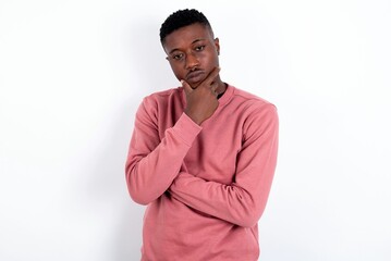 Thoughtful smiling young handsome man wearing pink sweater over white background keeps hand under chin, looks directly at camera, listens something with interest. Youth concept.