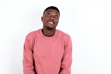 Portrait of mysterious young handsome man wearing pink sweater over white background looking up with enigmatic smile. Advertisement concept.