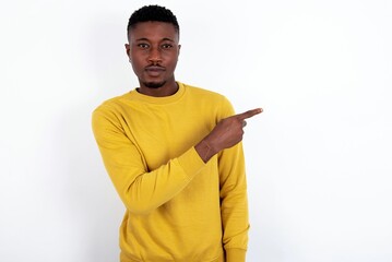 young handsome man wearing yellow sweater over white background smiling broadly at camera, pointing fingers away, showing something interesting and exciting.