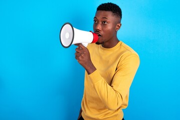 young handsome man wearing yellow T-shirt over blue background Through Megaphone with Available Copy Space