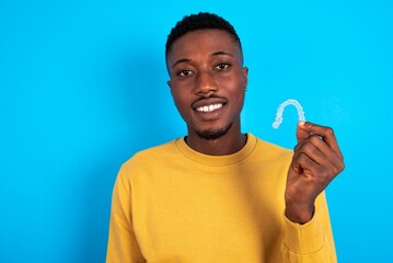 young handsome man wearing yellow T-shirt over blue background holding an invisible braces aligner, recommending this new treatment. Dental healthcare concept.