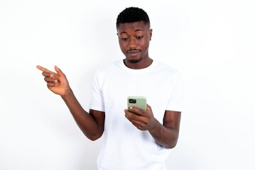 Smiling young handsome man wearing white T-shirt over white background pointing finger at blank space holding phone in one hand
