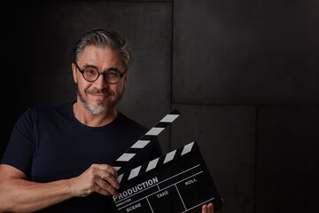 Happy man holding black clapper board. Movie director starting film. Copy space, dark background.