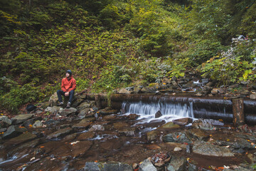 Active hiker in a red jacket sits next to a mountain river, resting after a challenging trail and...