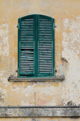 Fototapeta premium Window with closed green damaged wooden shutters, painted house wall with rough and cracked surface, no person, vertical format