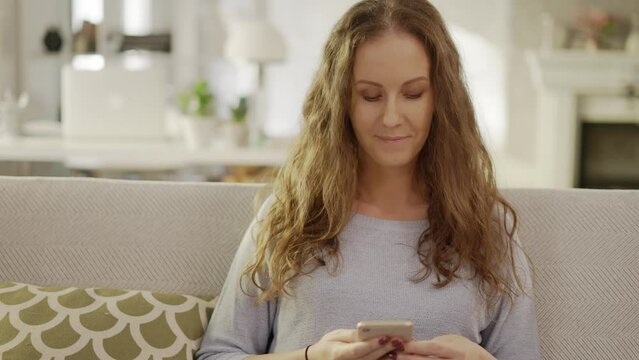 Woman at home, sitting on couch in living room, using phone, checking social media. Portrait of happy adult caucasian woman in 30s, smiling.