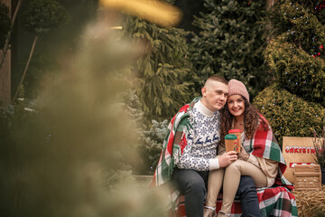 Beautiful young happy smiling couple wrapped in a Scottish blanket. Green Christmas trees on the...