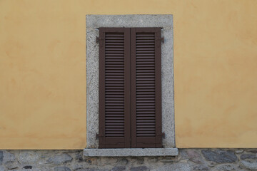 Fototapeta na wymiar Window with brown wooden shutters on a yellow cement wall and grey stone brick wall, grey frame around the window with windowsill, 