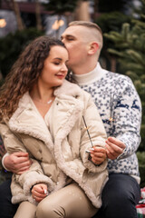 A beautiful young couple sitting near Christmas trees in white clothes walking near garlands. Happy man and woman, romance, selective focus, fun, love. White sweater, new year, street, outside