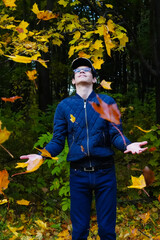 A teenage boy rejoices in the beauty of autumn and the falling maple leaves. Person looks up with a smile at the leaves falling around him and opens his palms to catch them, vertical photo