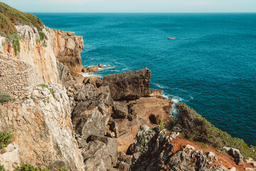 View from the top of a cliff of a rocky coastline with a reinforced slope above a beautiful teal seascape with a small boat bobbing on the ocean waves in the background; sunny day, Cascais, Portugal