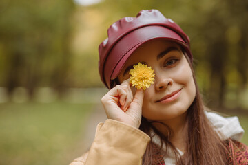 Portrait of smiling woman covering eye with yellow flower while hiding and looking at camera in nature in autumn 