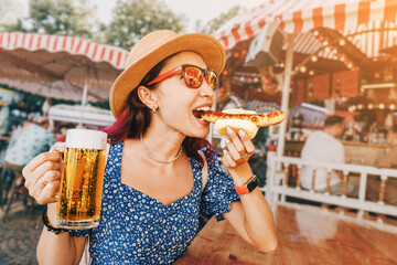 Happy girl drinking beer and eating traditional german bratwurst - hotdog at funfair and street food festival. National cuisine and biergarten concept - obrazy, fototapety, plakaty