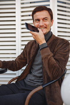 Smiling man recording voice message on smartphone and communicating on social media while sitting in outdoor cafe in city street 