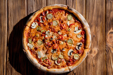 Appetizing pizza with hunting sausages, mozzarella cheese, pickled cucumbers, chili peppers on a tomato base on wood background