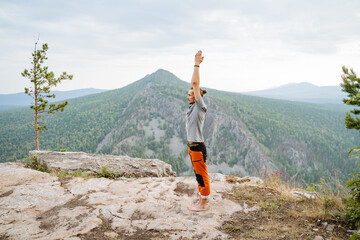 A young hipster guy standing on top of a mountain pulls his hands up, a man practices yoga in...