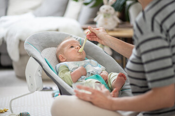 Mother spoon feeding her baby boy infant child in baby chair with fruit puree. Baby solid food...