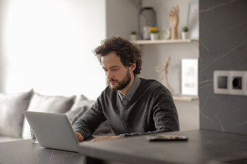 The man works at the leptop. The man is sitting at the table and typing on the computer. The guy works from home in a good mood. Work from home