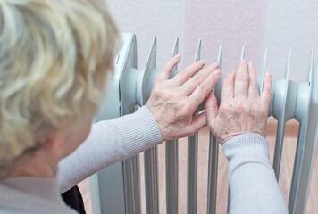 An elderly woman warms her hands on the radiator. Energy crisis, cold snap. Selective focus.