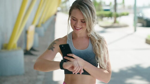 Young blonde woman wearing sportswear using smartphone and watch at street
