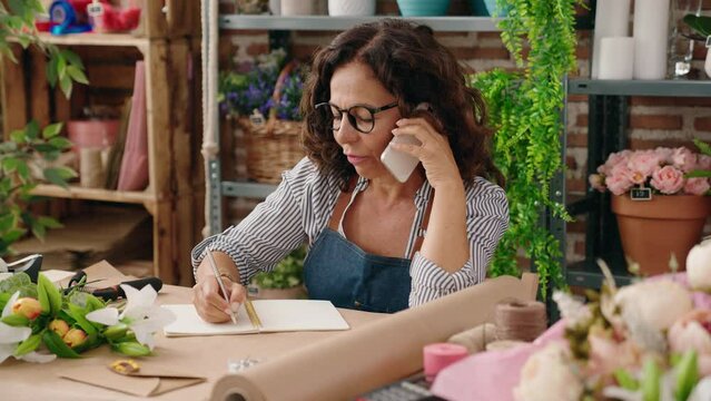 Middle age woman florist talking on smartphone writing on notebook at flower shop